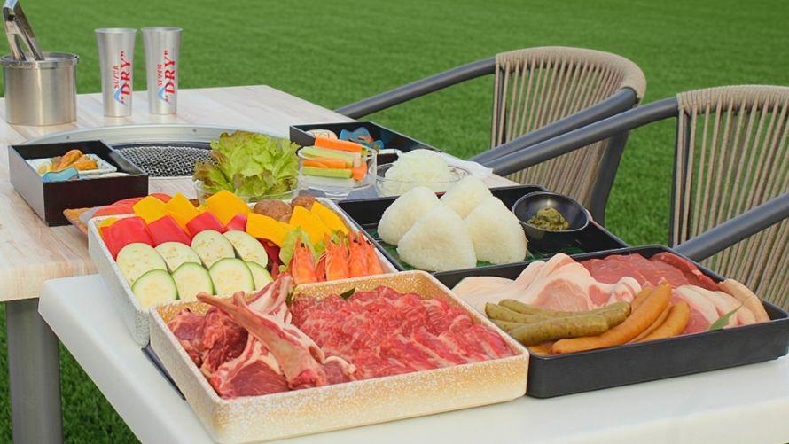 芝生の広がる屋外テラスで、テーブルの上に並べられた豪華なBBQ食材（牛肉、骨付き肉、ソーセージ、ズッキーニやパプリカなどの彩り豊かな野菜）。