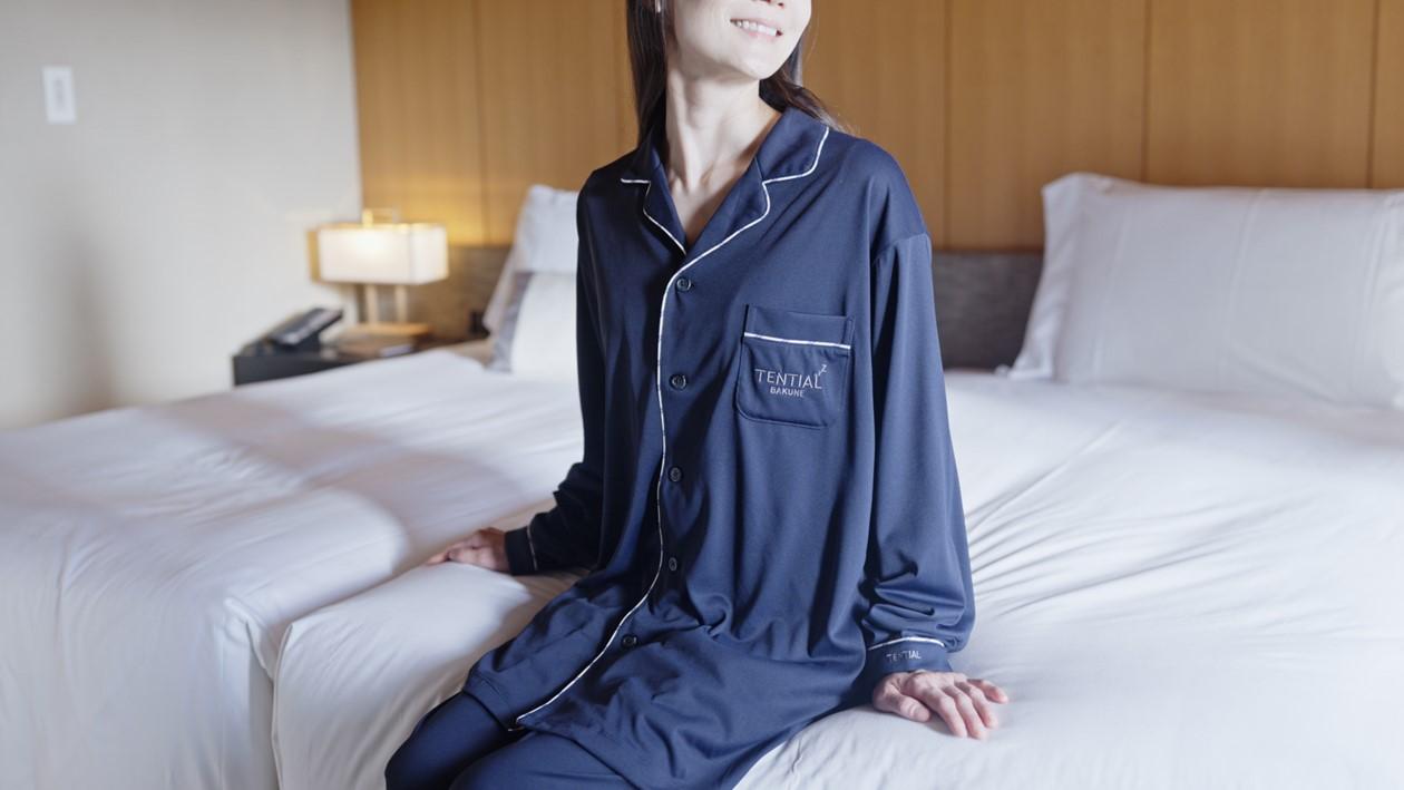 「BAKUNE」のパジャマを着た女性がベッドに腰かけてくつろいでいる。